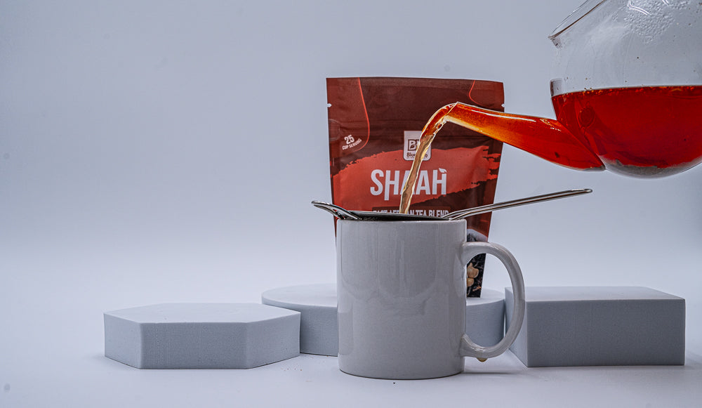 Picture of a red drink in a transparent glass kettle pouring the drink into a white cup with a Shaah package behind the white cup